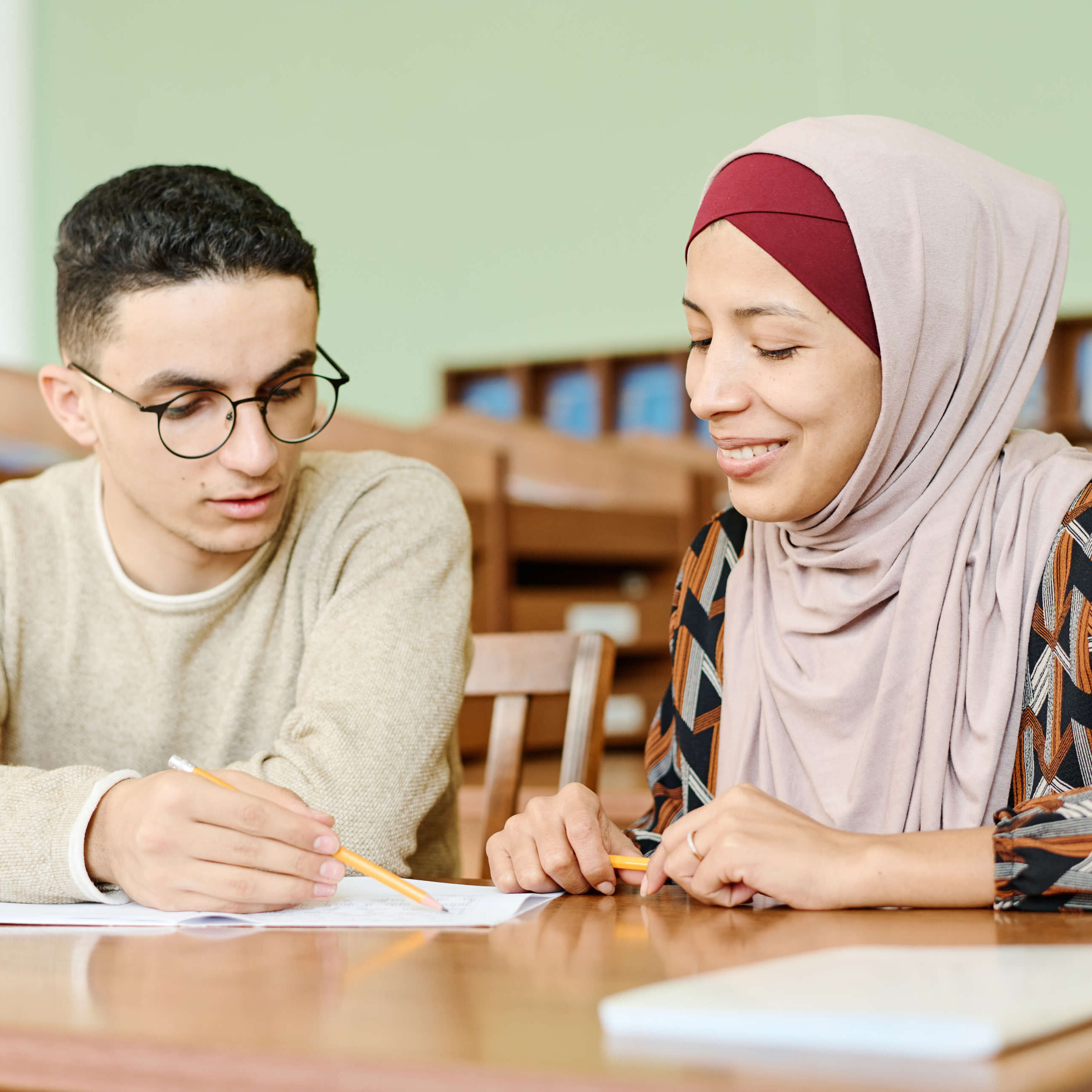 Young man and woman in hijab sitting at desk in classroom having lesson for immigrants working on writing task together